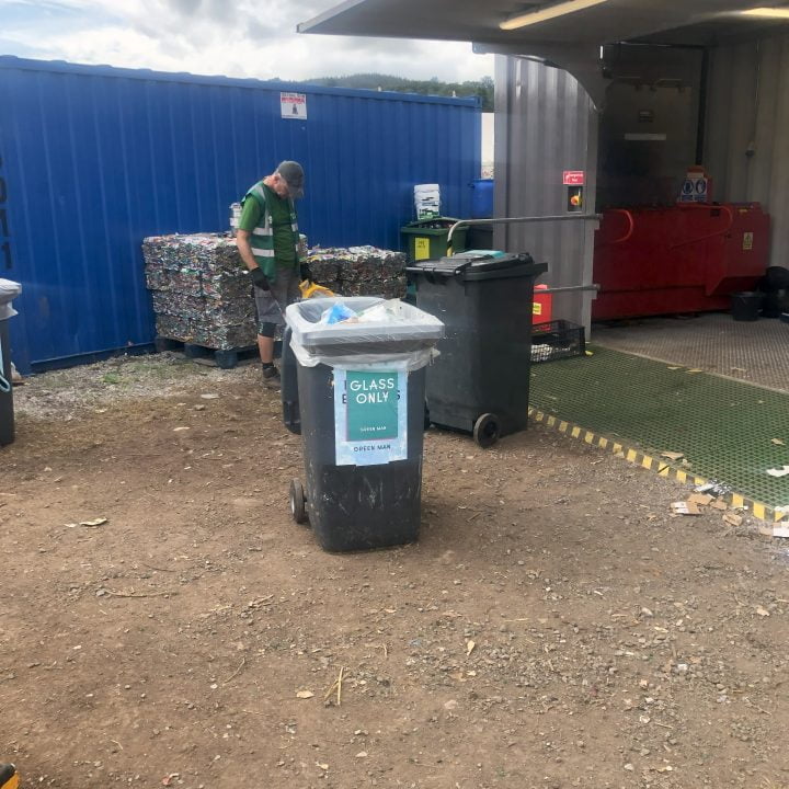 Workers prepare to sort out recycling. Wheelie bin marked up as glass. Bales of cans in the background.